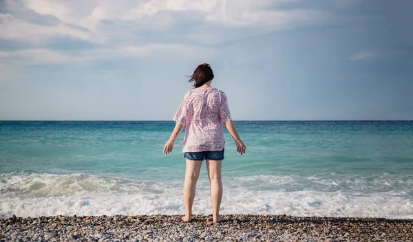 Jonge vrouw in korte broek en een shirt staat op een kiezelstrand en kijkt naar de golven van de Middellandse Zee, achteraanzicht, haar fladderen in de wind — Stockfoto