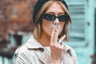 Uzun bej ceketli, beyaz çizmeli, siyah şapkalı ve sırt çantalı genç ve şık bir kadın. Mavi külüstür kapılar arkasında poz veriyor. Seçici odaklanma, hububat sokağı modası. yazarın tonu