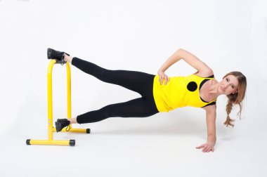 Sarışın kadın, beyaz arka planda yüksek sarı zemin parmaklıklarıyla egzersiz yapıyor. Siyah taytlı ve sarı tişörtlü bir kadın.