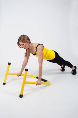 Sarışın kadın, beyaz arka planda yüksek sarı zemin parmaklıklarıyla egzersiz yapıyor. Siyah taytlı ve sarı tişörtlü bir kadın.