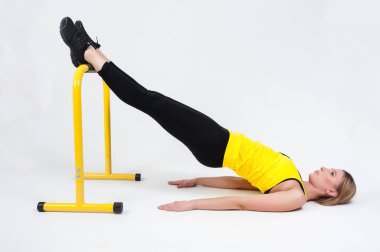 Sarışın kadın, beyaz arka planda yüksek sarı zemin parmaklıklarıyla egzersiz yapıyor. Siyah taytlı ve sarı tişörtlü bir kadın.