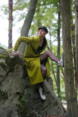 Yeşil paltolu şık bir genç kadın büyük beyaz çizmeler ve siyah taytlar bir ormanda bir taşın üzerinde oturuyor. Resim.