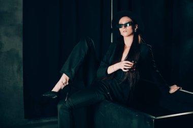 Siyah pantolonlu ve şapkalı bir kız siyah bir banyonun yanında poz veriyor. Elinde alkol olan bir bardak. Şık güneş gözlükleri.