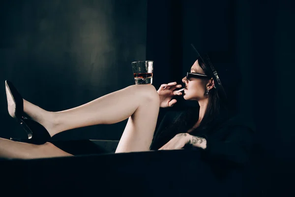 黒いジャケットの女の子と黒い風呂の近くにポーズをとってタバコを吸う帽子 手にアルコールとガラス スタイリッシュなサングラス 長い裸セクシー脚 — ストック写真