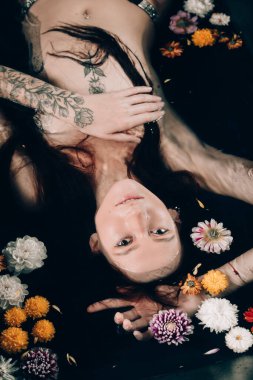 Küvette su ve çiçeklerle dövmeleri olan güzel, esmer bir kadın. Siyah bir banyoda sıradışı bir fotoğraf çekimi sırasında çıplak bir manken..