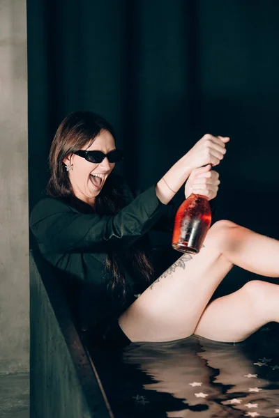 黒いバスルームの黒いジャケットの少女 バラのシャンパンワインのボトルを開きます スタイリッシュなサングラス 長い裸セクシー脚 — ストック写真