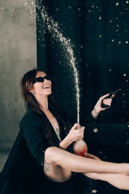 Siyah ceketli kız siyah bir banyoda poz veriyor. Elinde bir şişe gül şampanyası var. Şık güneş gözlükleri. Uzun çıplak seksi bacaklar. Şampanya damlaları