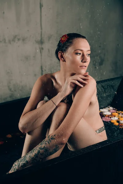 若いかわいいブルネットの女性は 水と花とお風呂で入れ墨 黒い風呂で珍しい写真撮影中の裸のモデル — ストック写真