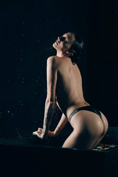 若いかわいいブルネットの女性は 水と花とお風呂で入れ墨 黒い風呂で珍しい写真撮影中の裸のモデル — ストック写真