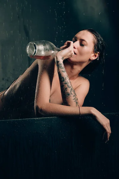 若いかなりブルネットの女性がバスルームにタトゥーを入れています 黒風呂で珍しい写真撮影中の裸のモデルワインを飲む — ストック写真