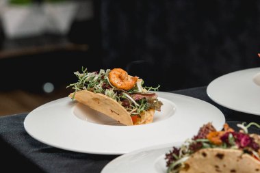 Salsa ve lahana salatalı baharatlı karidesli taco yemeye hazır bir masa. Masadaki beyaz tabakta birkaç taco var.