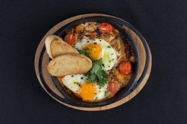 Sağlıklı kahvaltı shakshuka - kızarmış yumurta, soğan, dolmalık biber, domates ve baharatlar eski ahşap kalas üzerinde dökme demir güveç, dövüş açısı. Siyah masa örtüsü