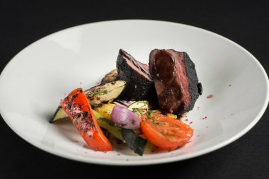 Küle bulanmış dana filetosu, siyah bir masa örtüsünde beyaz bir tabakta ızgara sebzeler..