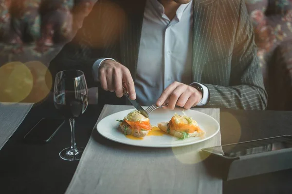 Молодой и привлекательный мужчина пользуется телефоном, в то время как вкусные яйца Бенедикт с слегка соленым лосося и соусом в закрытом ресторане — стоковое фото