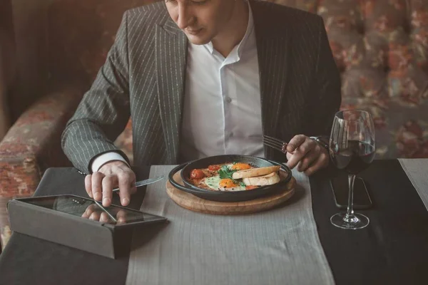 Человек использует свой планшет во время здорового завтрака шакшука - жареные яйца, помидоры и специи в чугунной стюпане на старой деревянной доске в закрытом ресторане. — стоковое фото