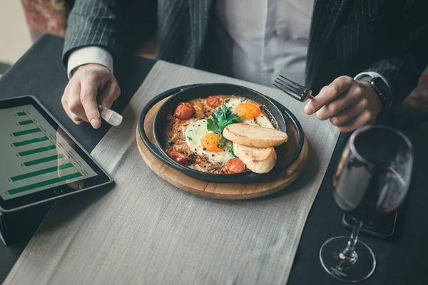 Człowiek używa tabletu podczas zdrowego śniadania shakshuka - smażone jajka, pomidory i przyprawy w stewpan żeliwny na starej drewnianej desce w restauracji wewnątrz. — Zdjęcie stockowe