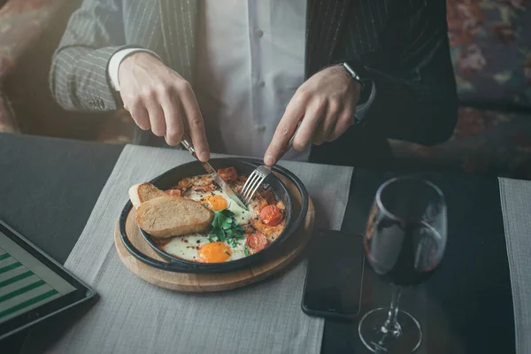Человек использует свой планшет во время здорового завтрака шакшука - жареные яйца, помидоры и специи в чугунной стюпане на старой деревянной доске в закрытом ресторане. — стоковое фото