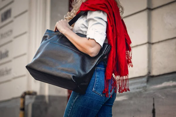 Fashionable woman posing in street with black leather bag. Model wearing trendy blue jeanse, scarf — Stock Photo, Image