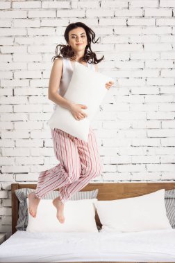 Beyaz yastıklı bir kız yatakta zıplıyor, beyaz tuğlalar arka planda. Rahat uyku kavramı. ortopedik yatak ve yastık