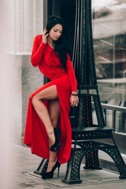 Kırmızı seksi elbiseli çekici genç kadın poz veriyor. Dekoratif kulenin yanında siyah saçlı baştan çıkarıcı bir kadının fotoğrafı. Seçici odak, film tahılları