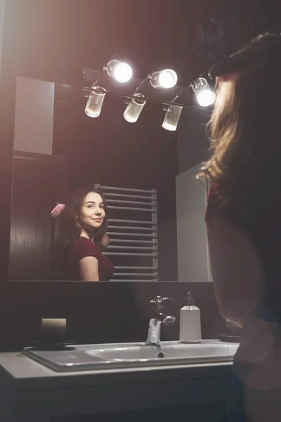 Νεαρή γυναίκα βουρτσίζει τα μαλλιά της μπροστά στον καθρέφτη του μπάνιου. — Φωτογραφία Αρχείου