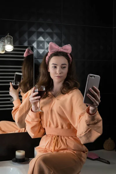Jolie jeune femme dans une salle de bain communiquant sur un smartphone et buvant du vin. Concept de détente et de loisirs. — Photo