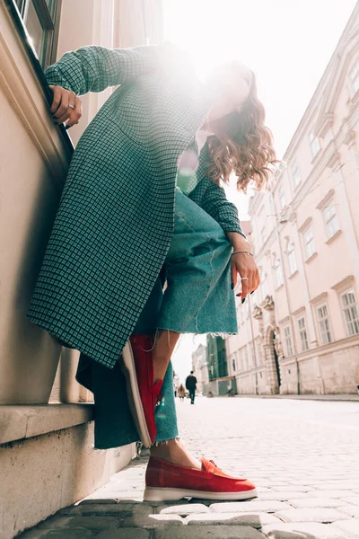 Woman in a red stylish suede loafer shoes, blue jeanse, white shirt and trendy checkered coat posing on the street. Pair of trendy female loafers shoes.