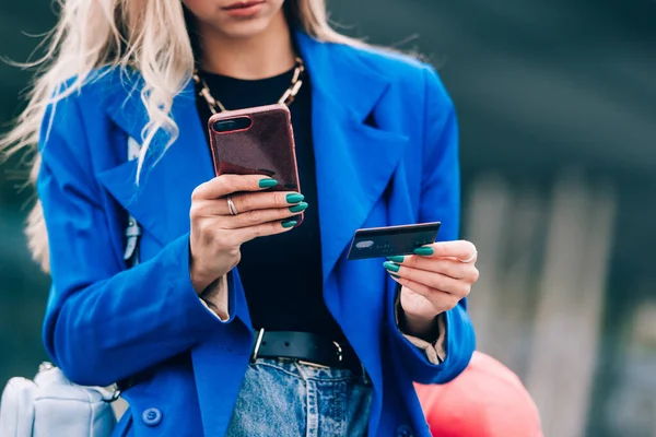 Comprador rubio de moda feliz con una chaqueta azul que paga en línea con una tarjeta de crédito y un teléfono inteligente en la calle — Foto de Stock