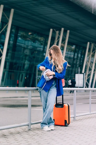 Portrait of a traveler woman standing with an orange suitcase near an airport. Young fashionable woman in a blue jeans and jacket, black shirt and white sneakers — Stock Photo, Image