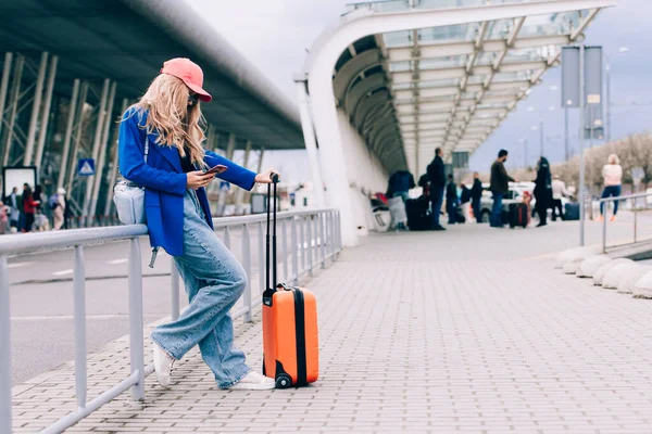 Retrato de uma mulher viajante de pé com uma mala laranja perto de um aeroporto. Mulher na moda jovem em uma calça azul e jaqueta, camisa preta e tênis brancos — Fotografia de Stock