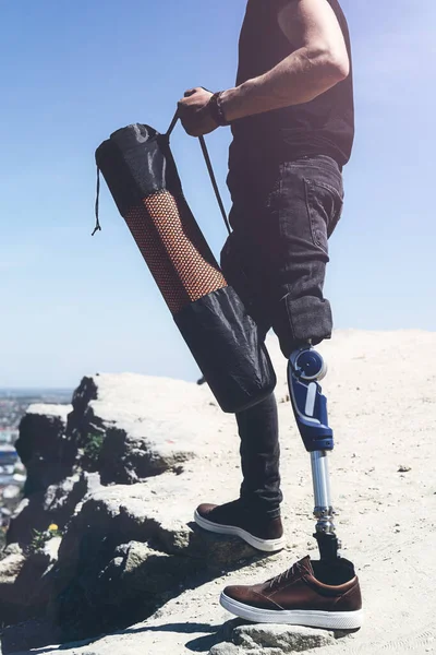 Um homem numa perna protética viaja pelas montanhas. Vestido com jeans pretos e uma camiseta, ele carregando tapete — Fotografia de Stock