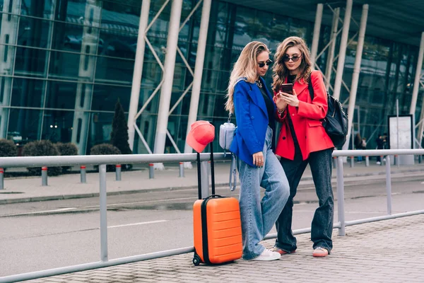 Dos chicas felices usando el vuelo de facturación de teléfonos inteligentes o el check-in en línea en el aeropuerto juntos, con equipaje. Viajes aéreos, vacaciones de verano —  Fotos de Stock