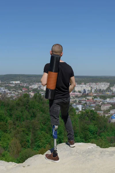 Um homem numa perna protética viaja pelas montanhas. Vestido com jeans pretos e uma camiseta, ele carregando tapete — Fotografia de Stock