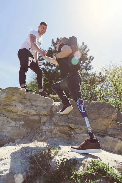 Um homem em uma perna protética viaja as montanhas com um amigo. Vestido com jeans pretos e uma camiseta, ele carregando tapete — Fotografia de Stock