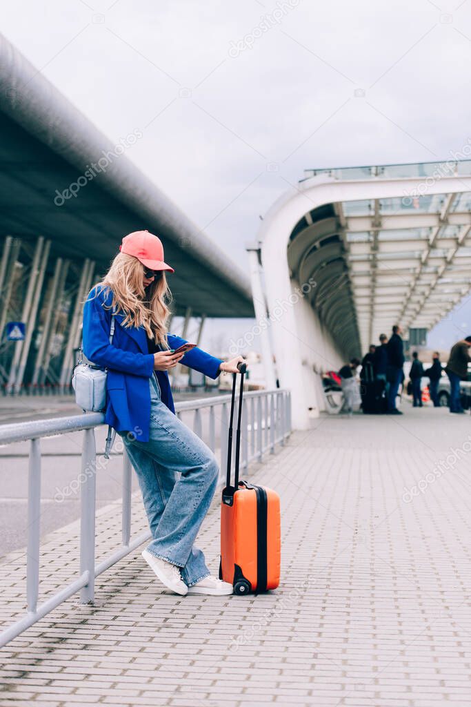 Portrait of a traveler woman standing with an orange suitcase near an airport. Young fashionable woman in a blue jeans and jacket, black shirt and white sneakers