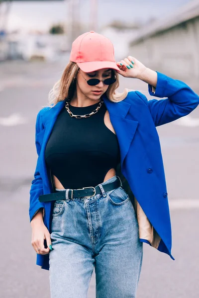 Joven sexy rubia hipster mujer posando en la calle. El uso de chaqueta elegante azul, jeans y sombrero de béisbol y gafas de sol. — Foto de Stock