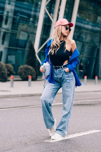 Jeune femme sexy blonde hipster posant dans la rue. Porter une veste bleue élégante, un jean, une casquette de baseball et des lunettes de soleil. — Photo