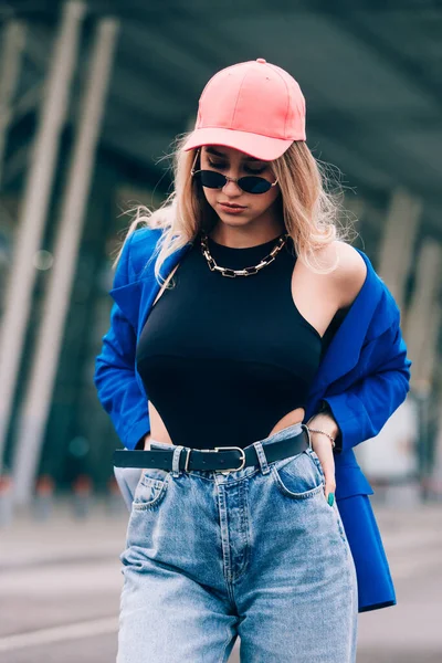 Jeune femme sexy blonde hipster posant dans la rue. Porter une veste bleue élégante, un jean, une casquette de baseball et des lunettes de soleil. — Photo