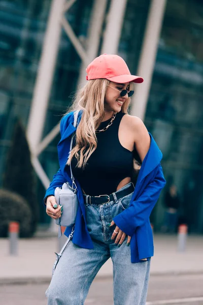Fiatal, szexi, szőke, hipszter nő pózol az utcán. Kék elegáns dzseki, farmer, baseball kalap és napszemüveg viselése. — Stock Fotó
