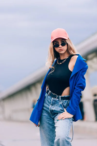 Jovem mulher hipster loira sexy posando na rua. Vestindo casaco azul elegante, jeans e chapéu de beisebol e óculos de sol. — Fotografia de Stock