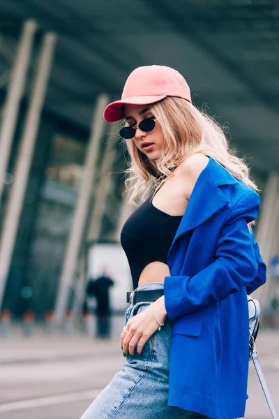 Молодая сексуальная блондинка-хипстер позирует на улице. Носит синий стильный пиджак, джинсы и бейсболку и солнцезащитные очки. — стоковое фото