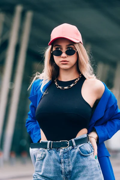 Молода сексуальна блондинка хіпстер позує на вулиці. Носіння синього стильного піджака, джинсів і бейсбольного капелюха і сонцезахисних окулярів . — стокове фото