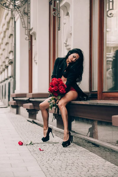 Κομψό όμορφη μελαχρινή γυναίκα σε κόκκινο σορτς και μαύρο παλτό κορίτσι που θέτουν κάθεται στο περβάζι του παραθύρου με ένα μεγάλο μπουκέτο κόκκινα τριαντάφυλλα. Γενέθλια. Ημέρα του Αγίου Βαλεντίνου. Επιλογή εστίασης, filmgrain — Φωτογραφία Αρχείου