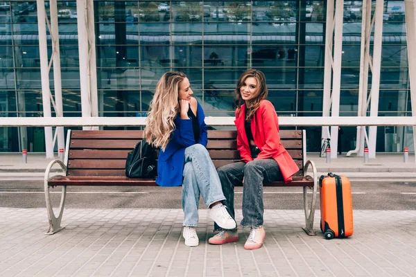 Dos chicas felices sentadas en un banco cerca del aeropuerto, con equipaje. Viajes aéreos, vacaciones de verano —  Fotos de Stock