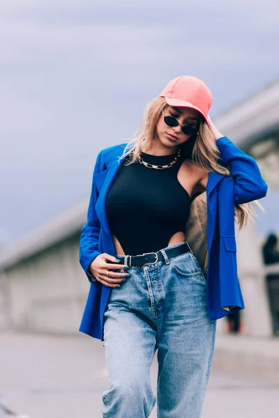 Молода сексуальна блондинка хіпстер позує на вулиці. Носіння синього стильного піджака, джинсів і бейсбольного капелюха і сонцезахисних окулярів . — стокове фото