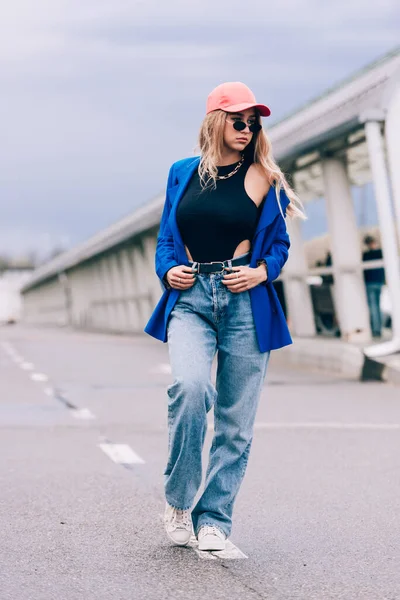 年轻性感金发嬉皮士女人在街上摆姿势。穿着蓝色时髦夹克、牛仔裤、棒球帽和太阳镜. — 图库照片
