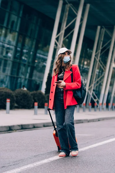 Portrait of a traveler woman in a mask walking with an orange suitcase near an airport. Young fashionable woman in a blue jeans and jacket — Stock Photo, Image