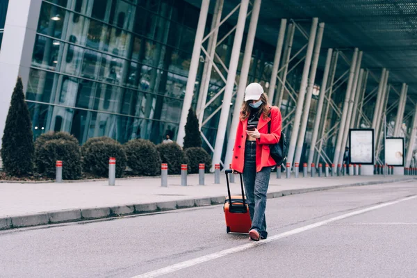 Retrato de uma mulher viajante em uma máscara andando com uma mala laranja perto de um aeroporto. Mulher na moda jovem em uma calça azul e jaqueta — Fotografia de Stock
