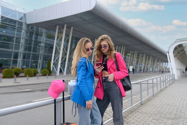 Duas meninas felizes usando smartphone verificando voo ou check-in on-line no aeroporto juntos, com bagagem. Viagens aéreas, férias de verão — Fotografia de Stock