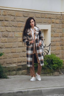 Kareli uzun palto, bej pantolon ve beyaz bluz giyen güzel ve modaya uygun genç bir kadının yakın plan portresi. Şehir caddesinde poz veren kadın.